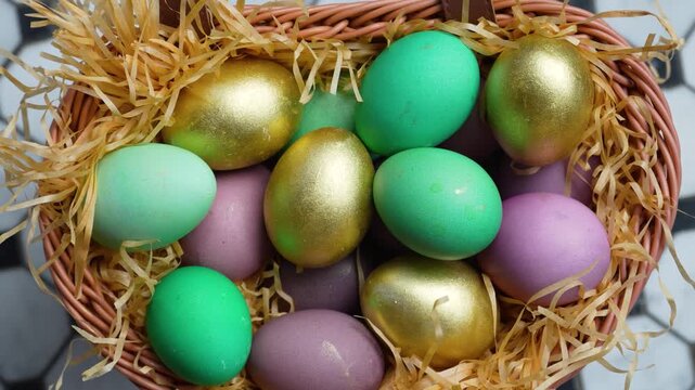 Colorful Easter Eggs in Wicker Basket with Straw. A wicker basket filled with decorative Easter eggs in gold, green, and purple hues nesting on wood wool
