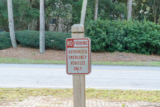 "No Parking Authorized Emergency Vehicles Only" sign on a wooden post by a paved road. White sign with red lettering placed near a curb with green landscaping, bushes, and trees in the background.