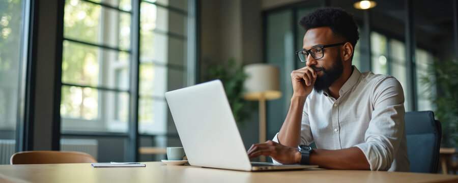 Black man in glasses typing on laptop computer. He wears light shirt, sits at table in office. Man looks thoughtful, holding hand near mouth, thinking about work.
