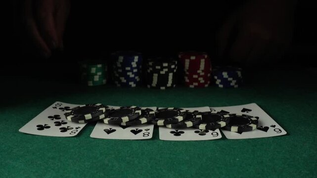 Hand positions black poker chips on top of playing cards during a card game on a green felt table, with stacks of colorful poker chips visible in the background