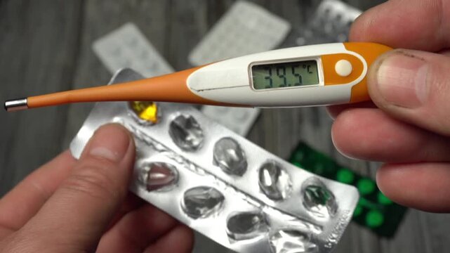Person checks temperature with digital thermometer, examines blister pack of capsules, and prepares to take medication in a well-lit indoor setting