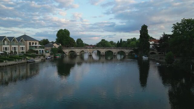 A serene river flows beneath an arched stone bridge, lined with trees and modern houses, under a cloudy sky.
