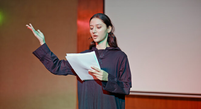 Indian young adult gen z female lady standing wear outfit dress hold script paper do play scene practice indoor hall stage. Teen age girl read story line talk speak day time college campus room place