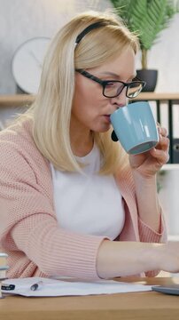 Remote work concept where good-looking confident successful blond businesswoman sitting in front of computer in cozy home office and making notes into report when reading datas from laptop screen