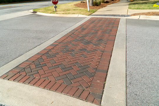 Vibrant herringbone brick paver crosswalk featuring red and dark grey tones, providing a safe pedestrian path across a textured asphalt road, bordered by smooth concrete curbs in a sunny suburban sett