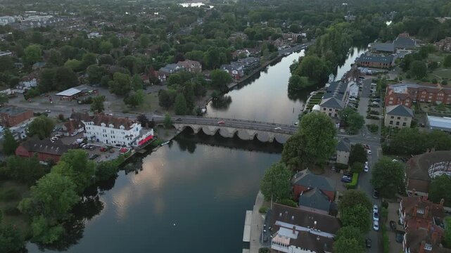 Aerial view of a town with a river and a stone bridge. Buildings line the riverbanks, surrounded by lush trees.