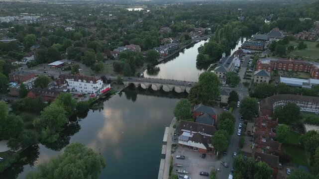 Drone footage shows a scenic riverbend with a bridge, riverside buildings, and lush greenery, capturing a tranquil evening atmosphere.