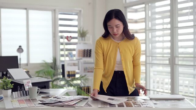 Asian young adult woman designer reviewing color swatches material samples documents in modern bright office workspace professional project planning