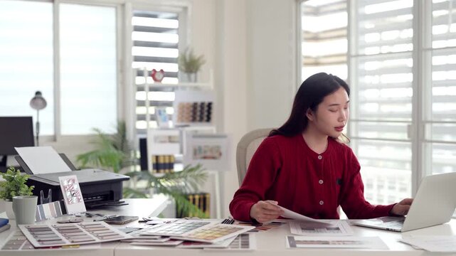 Asian adult female designer working creative project planning using laptop computer color swatches printer modern home office environment