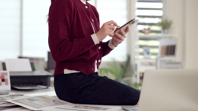 Asian adult woman uses smartphone tapping screen checking messages browsing internet office desk workspace productivity digital communication