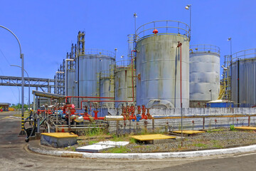 Obraz premium A series of large cylindrical storage tanks and complex piping systems at a petrochemical refinery under a clear blue sky. The facility features prominent red valves and metallic conduits used for the