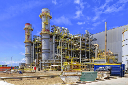 A wide-angle view of a complex industrial power plant featuring tall exhaust chimneys, intricate pipe systems, and steel framework structures. The facility is set against a vibrant blue sky with scatt