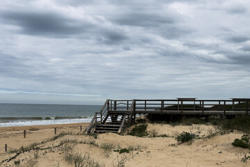 Fototapeta premium Sturdy wooden stairs rise from the sandy beach, surrounded by tall grasses, leading to the top of the dune under a cloudy afternoon sky. The scene is calm and inviting.