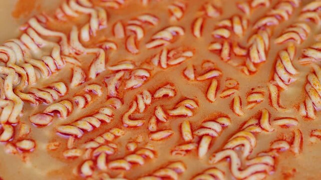 Noodles in broth. Macro shot of noodles boiling in flavorful orange spice broth. Detailed footage of curly noodles releasing starch in spicy orange broth with bubbles