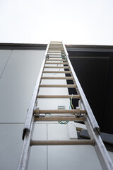 An aluminium ladder is leaning on the house roof, using for climbing ascent up to roof top for building activity susch as installing the solar cell. Close-up and selective focus at ladder's part. © Nattawit