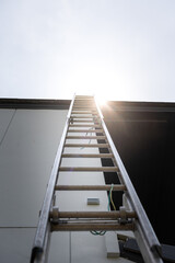 An aluminium ladder is leaning on the house roof, using for climbing ascent up to roof top for building activity susch as installing the solar cell. Close-up and selective focus at ladder's part. © Nattawit