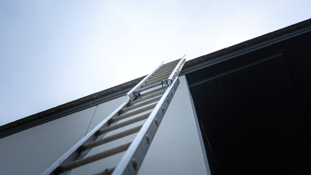 An aluminium ladder is leaning on the house roof, using for climbing ascent up to roof top for building activity susch as installing the solar cell. Close-up and selective focus at ladder's part.