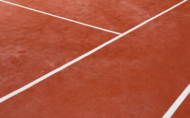 Fototapeta premium A clay court, marked with bright white lines, awaits the next set. Tennis balls scattered across the surface hint at a recent practice. Greenery surrounds the outdoor space.