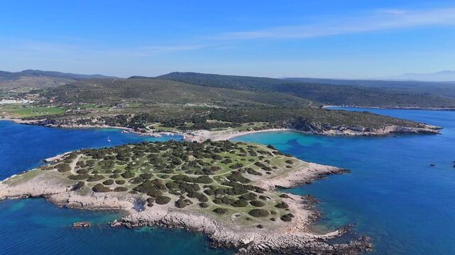 A high-altitude aerial view showcasing a rugged Mediterranean island connected to a mainland bay by a narrow, sandy tombolo.