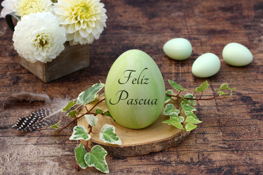 Un huevo de Pascua verde con la inscripci&oacute;n "Feliz Pascua" en un disco de madera con zarcillos de hiedra, junto a &eacute;l flores blancas en una caja de madera y tres huevos verdes m&aacute;s sobre una mesa de mad