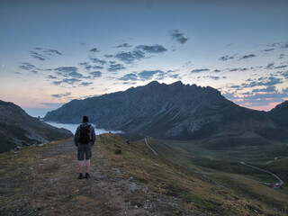 Fototapeta premium Hiker watching mountain sunset over fog - Rear view of a hiker with a backpack standing on a ridge, gazing at a vast mountain panorama over a misty valley at dusk or dawn.