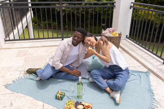 Diverse couple sitting on light blue blanket on rooftop patio sharing fruit and wine