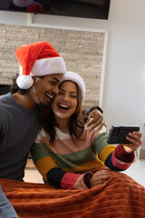 Fototapeta premium Diverse couple laughing while taking selfie on couch at home with smartphone and Santa hats