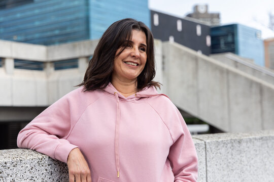 Middle-aged woman leaning on speckled parapet on concrete terrace wearing light pink hoodie