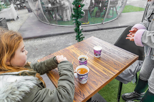A high angle shot of a young girl and her mother sitting at a wooden table in an outdoor cafe with paper cups of hot tea and coffee in a sunny autumn park with bokeh background and copy space