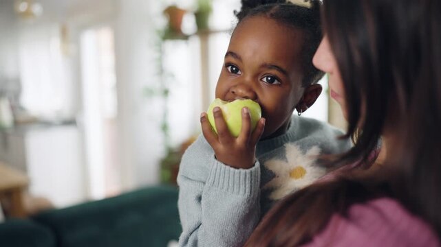 Adorable little girl sitting on her mother's lap, eating a fresh green apple. A tender moment of healthy snacking and family love at home