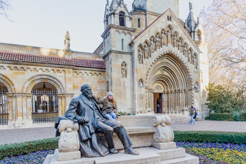A happy young girl in a winter hat sitting on a stone bench next to the bronze statue of architect Ignác Alpár in front of the historic stone facade of Ják Chapel in Budapest City Park today © Igor