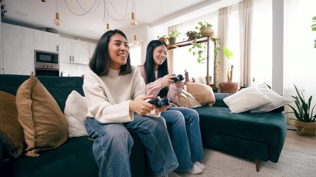 Happy asian women having fun playing video games with a controller. An excited lesbian couple sitting on the couch in a modern living room