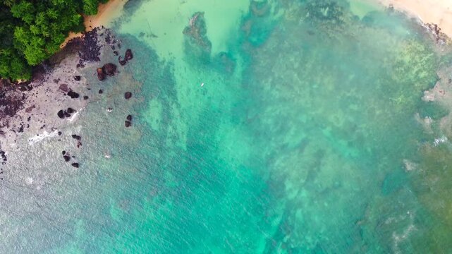 An aerial top-down video of clear turquoise water meeting a dark rocky shoreline and lush tropical forest on S&atilde;o Tom&eacute; Island, S&atilde;o Tom&eacute; and Pr&iacute;ncipe