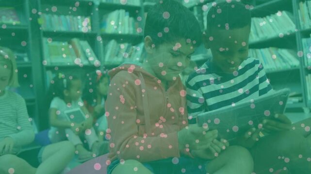 Two boys leaning in and tapping tablet in school library, sharing books while pink dots overlaying