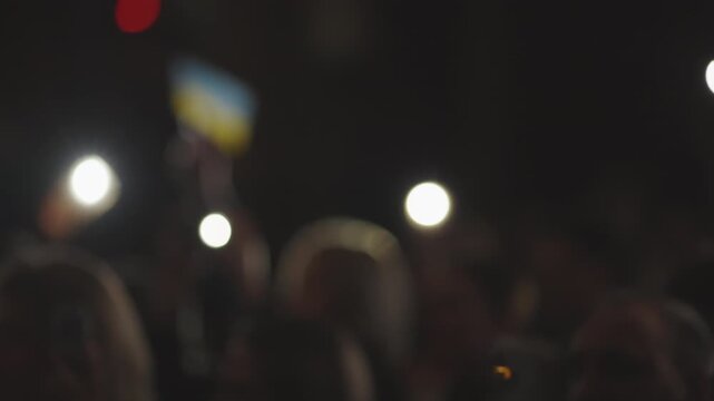A close-up of a hand waving a smartphone with its flashlight on in a dark, crowded setting. Many other phone flashlights create a vibrant, energetic concert atmosphere
