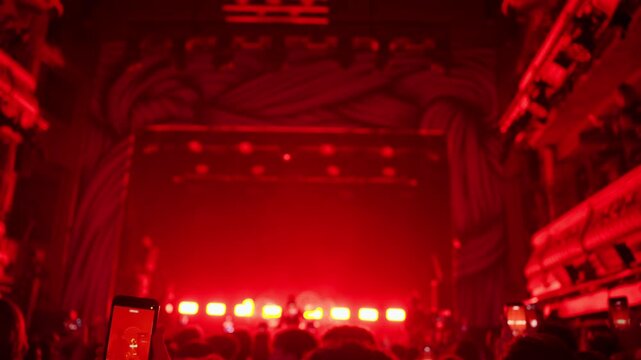 An enthusiastic crowd raises their hands and makes rock gestures at a vibrant live music concert bathed in intense red stage lighting. The atmosphere is energetic and exciting