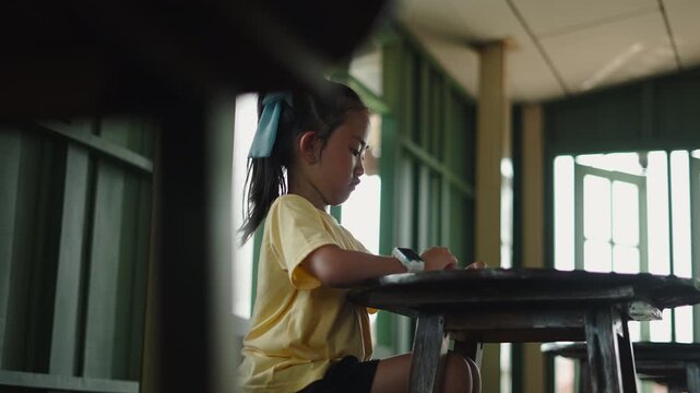 Contrapicated view of a little girl with a blue bow focuses on writing on a wooden table in a room with green windows and sunlight