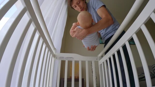 Dad gently puts his 6 months old baby boy into a crib at home, creating a calm and loving bedtime routine. Slowmotion video.
