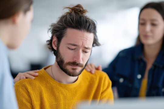 Young caucasian male adult upset supported by coworkers comforting gesture at office desk