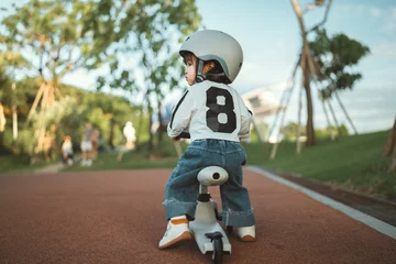 Rear view - cute Asian toddler girl wearing a safety helmet while learning to ride a balance bike at a public park during a sunny afternoon, representing early childhood development, outdoor © oatawa