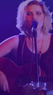 Vertical video: Music artist leaning into mic singing and playing guitar under blue lights to play