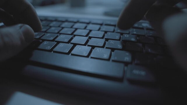 Close up on Hebrew and English computer keyboard as hands type in dark office. Hacker or spy using keyboard clicking on buttons with Hebrew letters. Extreme close up. Technology mystery anonymous