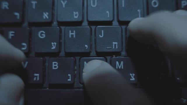 Top shot on fingers typing on hebrew computer keyboard keys in dark room. Person working on computer writing document using keyboard with Hebrew letters. Hacker or spy coding. Extreme close up