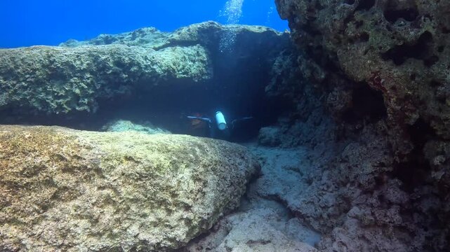 Scuba diver swimming into underwater cave. Diver exploring the tunnel in the rocky seascape, vivid blue ocean. Underwater cave and swimming scuba diver. Underwater exploration, travel footage. 