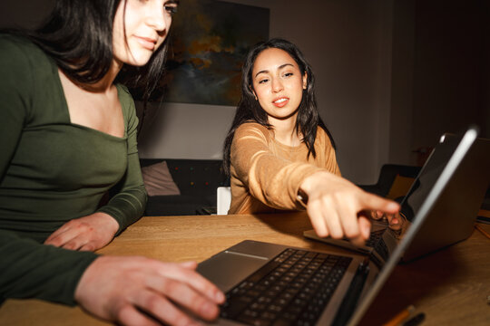 Two young women collaborate on a project using laptops at a wooden table, one pointing at the screen to explain something.