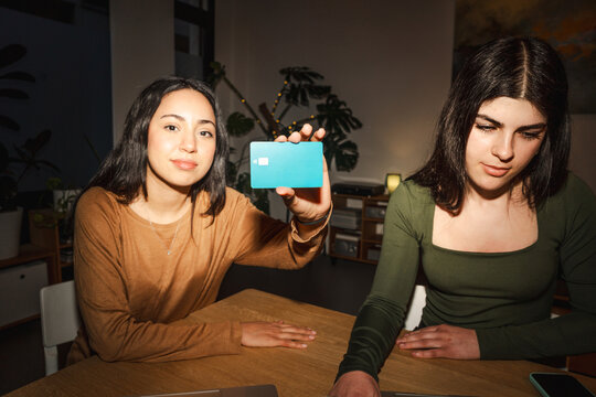 Two young women are at a table, one holding up a debit card, the other looking down at a laptop.