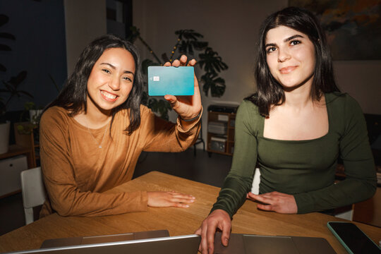 Two young women are sitting at a table, one holding up a debit card, suggesting financial transactions or banking.
