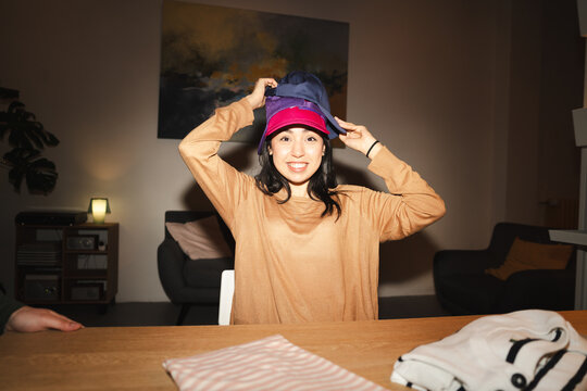 A young woman smiles as she stacks multiple baseball caps on her head, enjoying a moment of playful fun indoors.