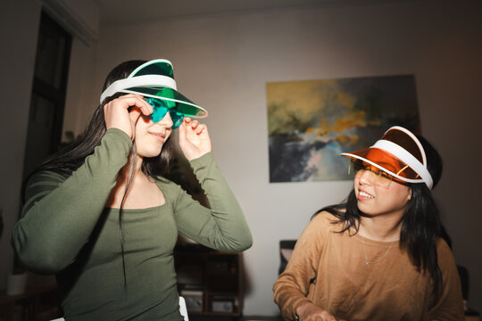 Two young women wearing colorful visors and sunglasses are having fun indoors, adjusting their accessories.