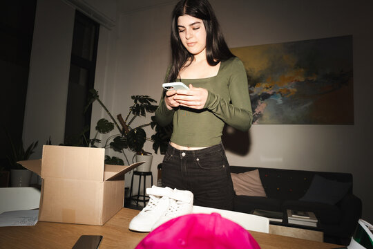 A young woman uses her phone while standing next to an open box with new shoes and a pink hat on a wooden table.